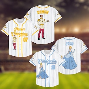 May include: White baseball jerseys with pinstripes, featuring cartoon illustrations of Prince Charming and Cinderella. The jerseys have gold and blue accents, with names and numbers on the back and front.