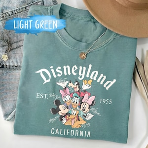 Puede incluir: Camiseta verde claro con la palabra "Disneyland" en escritura blanca, con personajes de dibujos animados. La camiseta incluye el texto "EST. 1955 CALIFORNIA". Tambi&eacute;n se ven un collar dorado y un sombrero marr&oacute;n.