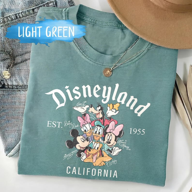 Puede incluir: Camiseta verde claro con la palabra "Disneyland" en escritura blanca, con personajes de dibujos animados. La camiseta incluye el texto "EST. 1955 CALIFORNIA". Tambi&eacute;n se ven un collar dorado y un sombrero marr&oacute;n.
