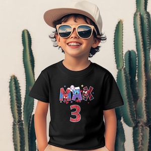 Pode incluir: Uma camiseta preta com um gr&aacute;fico de personagens do Homem-Aranha que soletram o nome "MAX" e o n&uacute;mero "3" em vermelho. A camiseta est&aacute; sendo usada por um menino pequeno que est&aacute; usando um bon&eacute; de beisebol branco e &oacute;culos de sol.