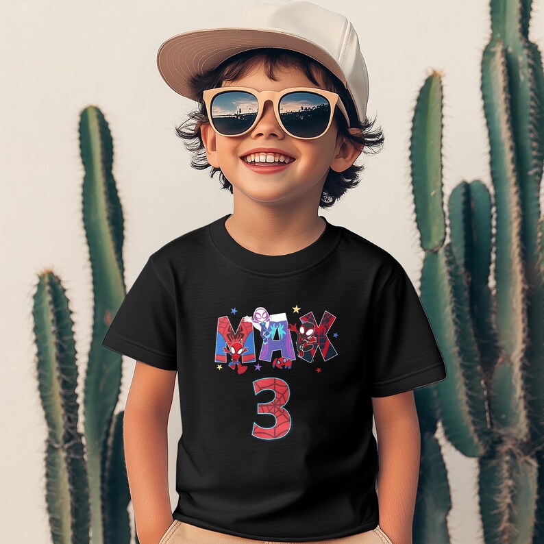 Pode incluir: Uma camiseta preta com um gr&aacute;fico de personagens do Homem-Aranha que soletram o nome "MAX" e o n&uacute;mero "3" em vermelho. A camiseta est&aacute; sendo usada por um menino pequeno que est&aacute; usando um bon&eacute; de beisebol branco e &oacute;culos de sol.