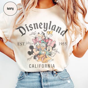 Puede incluir: Camiseta de color marfil con la palabra "Disneyland" en una fuente vintage y personajes de Disney. La camiseta incluye el texto "EST. 1955 CALIFORNIA" y las firmas de los personajes. La camiseta es de color crema claro.
