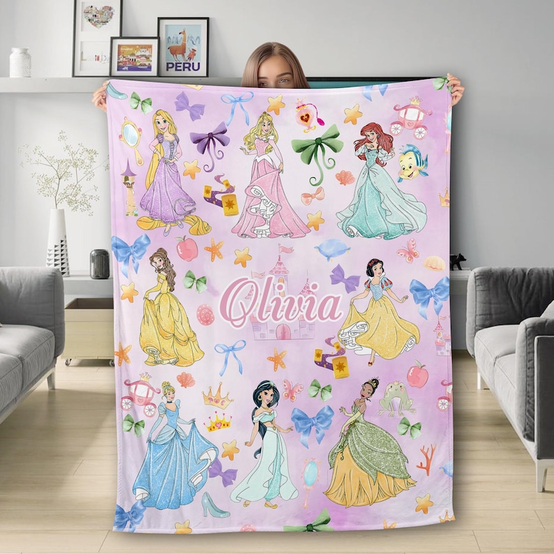 May include: A pink fleece blanket with a pattern of Disney princesses, including Cinderella, Snow White, Ariel, Belle, and Jasmine. The blanket features a pink background with gold stars, bows, and a castle. The name "Olivia" is written in gold letters on the blanket.