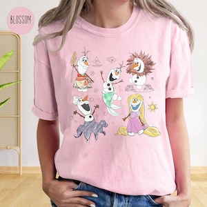 Pode incluir: Camiseta rosa com uma ilustra&ccedil;&atilde;o de desenho animado de cinco bonecos de neve. Os bonecos de neve est&atilde;o vestidos com roupas diferentes, incluindo uma camisa havaiana, uma cauda de sereia e um vestido roxo. Os bonecos de neve est&atilde;o cercados por bolhas e um sol.
