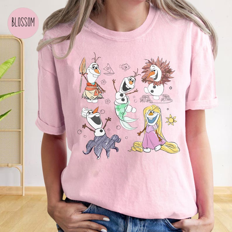 Pode incluir: Camiseta rosa com uma ilustra&ccedil;&atilde;o de desenho animado de cinco bonecos de neve. Os bonecos de neve est&atilde;o vestidos com roupas diferentes, incluindo uma camisa havaiana, uma cauda de sereia e um vestido roxo. Os bonecos de neve est&atilde;o cercados por bolhas e um sol.
