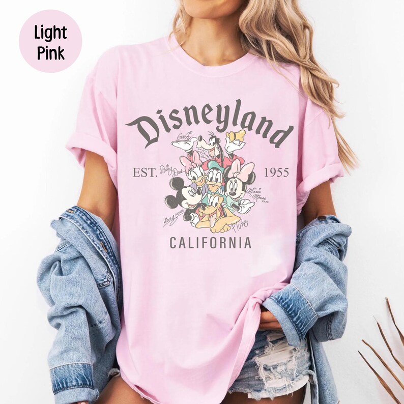 Puede incluir: Camiseta rosa claro con el logotipo de Disneyland y Mickey Mouse, Minnie Mouse, Donald, Goofy y Pluto. La camiseta incluye el texto "EST. 1955 CALIFORNIA". La camiseta es de color rosa claro.
