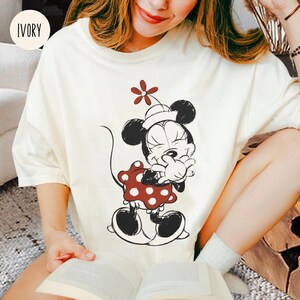 Op de afbeelding: Ivoor t-shirt met een zwart-wit illustratie van een stripfiguur. Het personage draagt een rode jurk met witte stippen en een witte hoed met een rode bloem. Het t-shirt heeft korte mouwen.