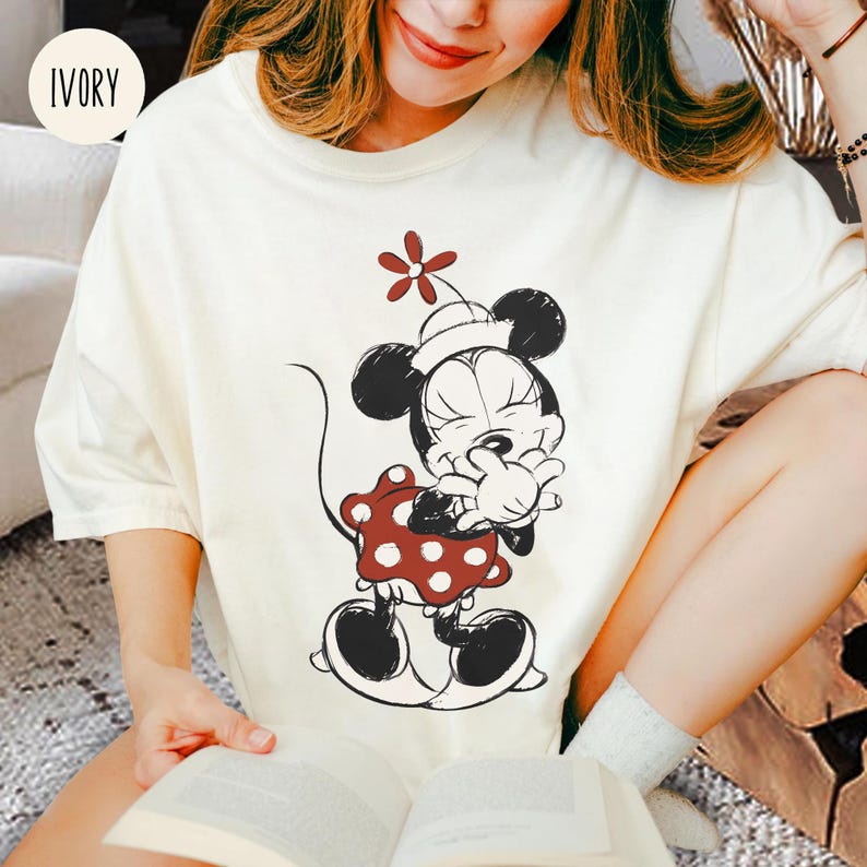 Op de afbeelding: Ivoor t-shirt met een zwart-wit illustratie van een stripfiguur. Het personage draagt een rode jurk met witte stippen en een witte hoed met een rode bloem. Het t-shirt heeft korte mouwen.