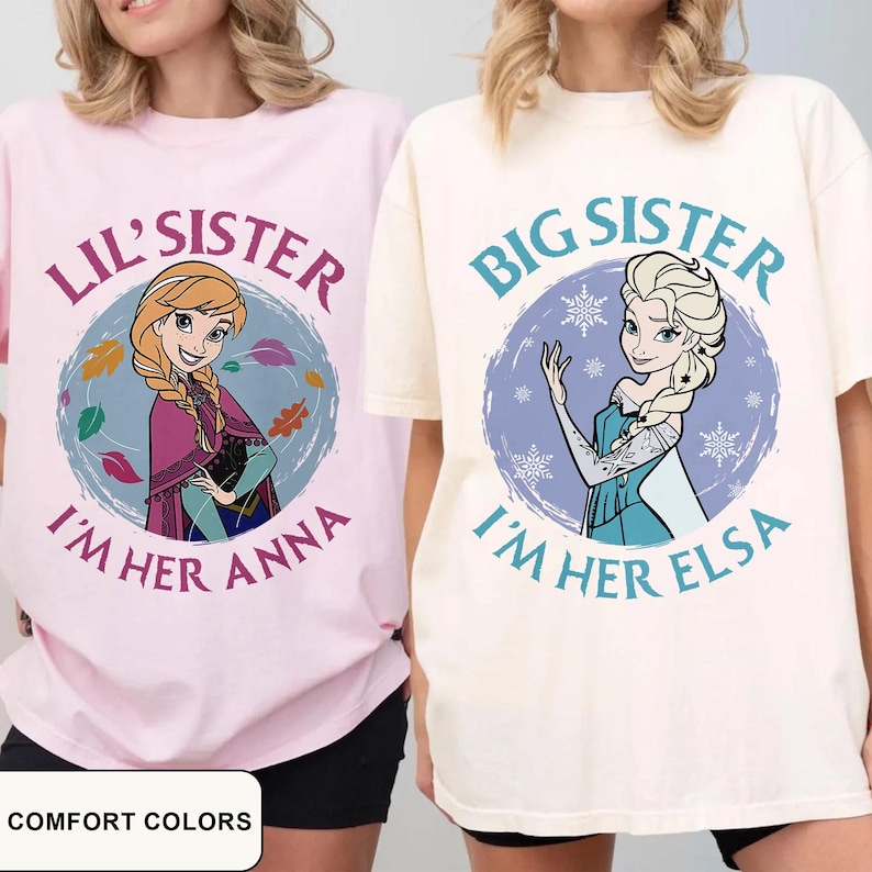 May include: Two t-shirts, one pink and one cream, featuring cartoon characters. The pink shirt says "Lil' Sister I'm Her Anna" with an Anna graphic. The cream shirt says "Big Sister I'm Her Elsa" with an Elsa graphic. The text "Comfort Colors" is at the bottom.
