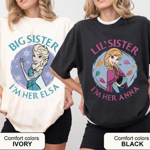 May include: Two t-shirts, one ivory and one black, featuring Disney's Frozen characters. The ivory shirt says "Big Sister, I'm Her Elsa" with Elsa's image. The black shirt says "Lil' Sister, I'm Her Anna" with Anna's image.