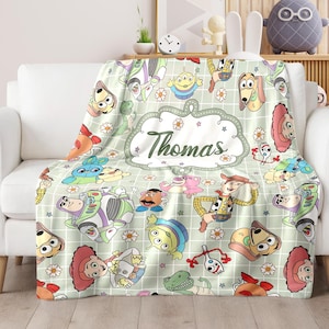 May include: A green and white fleece blanket with a grid pattern and a variety of Toy Story characters, including Woody, Buzz Lightyear, and the aliens. The blanket has a personalized name, "Thomas", in a white oval with green stars and flowers.