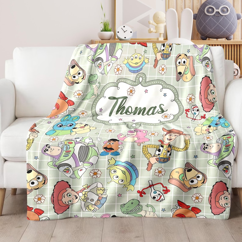 May include: A green and white fleece blanket with a grid pattern and a variety of Toy Story characters, including Woody, Buzz Lightyear, and the aliens. The blanket has a personalized name, "Thomas", in a white oval with green stars and flowers.