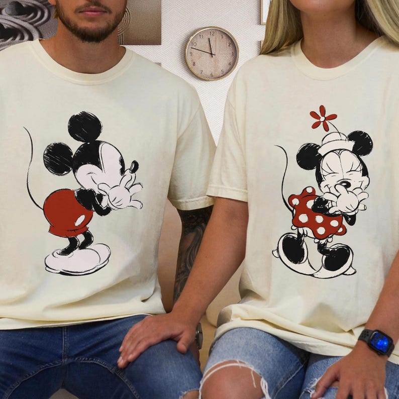 Op de afbeelding: Twee cr&egrave;mekleurige T-shirts met illustraties van Mickey en Minnie Mouse. Mickey draagt een rode korte broek en Minnie een rode jurk met stippen. Beide personages staan in een speelse pose. De shirts worden door twee mensen gedragen, met een blauwe jeans.