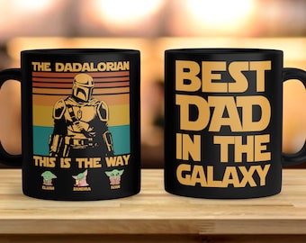May include: Two black coffee mugs. One mug features a retro-style illustration of a Mandalorian character with the text "The Dadalorian" and "This is the Way". The other mug has the text "Best Dad in the Galaxy".