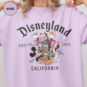 Puede incluir: Una camiseta lila con la palabra "Disneyland" en escritura negra, y el texto "EST. 1955 CALIFORNIA". La camiseta presenta un gr&aacute;fico de personajes de Disney como Mickey Mouse, Minnie Mouse y Goofy.