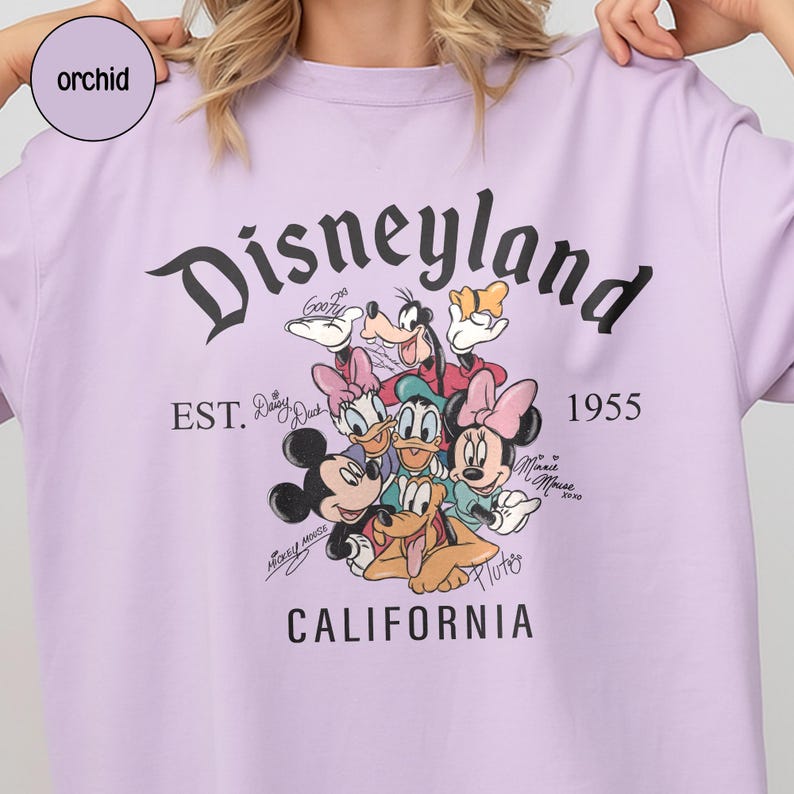 Puede incluir: Una camiseta lila con la palabra "Disneyland" en escritura negra, y el texto "EST. 1955 CALIFORNIA". La camiseta presenta un gr&aacute;fico de personajes de Disney como Mickey Mouse, Minnie Mouse y Goofy.