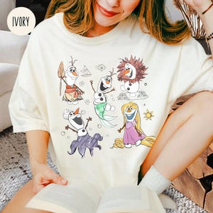 Pode incluir: Uma camiseta branca com um desenho de desenho animado de Olaf, Elsa, Anna e Rapunzel. Os personagens s&atilde;o desenhados em um estilo simples com cores brilhantes. A camiseta &eacute; de cor marfim claro.