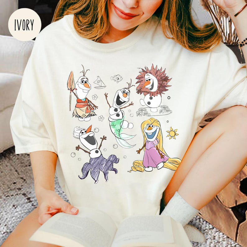 Pode incluir: Uma camiseta branca com um desenho de desenho animado de Olaf, Elsa, Anna e Rapunzel. Os personagens s&atilde;o desenhados em um estilo simples com cores brilhantes. A camiseta &eacute; de cor marfim claro.
