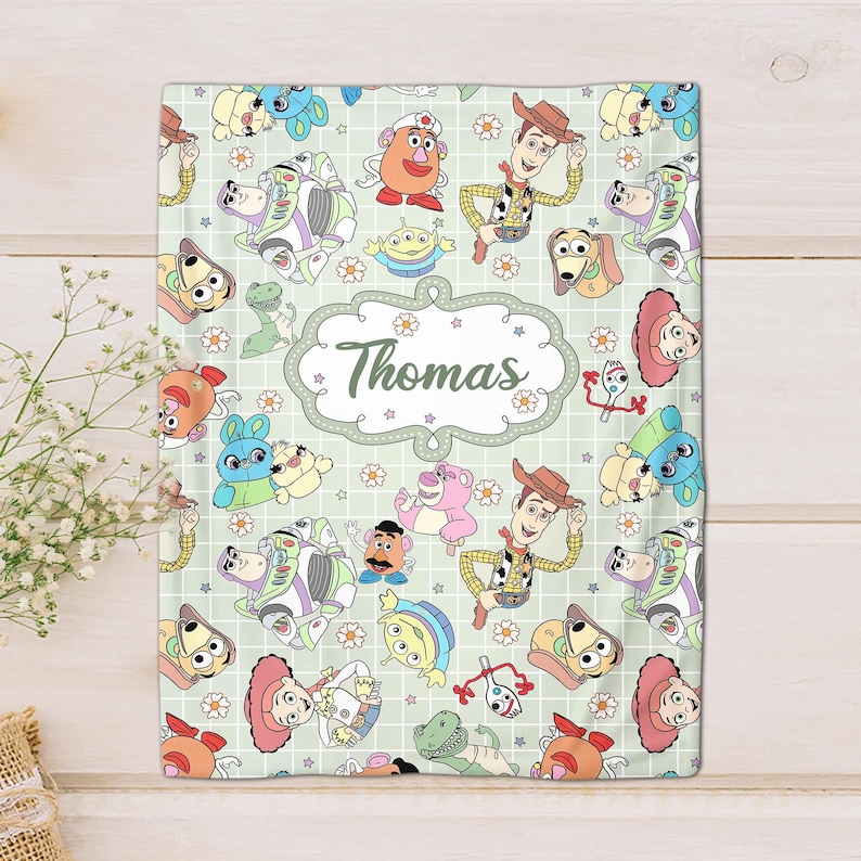 May include: A green and white blanket with a repeating pattern of Toy Story characters, including Woody, Buzz Lightyear, and Mr. Potato Head. The blanket has a personalized name, "Thomas", in a decorative font.