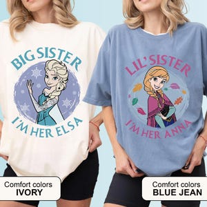 May include: Two t-shirts, one ivory and one blue jean, featuring cartoon images of Elsa and Anna from Frozen. The ivory shirt says "Big Sister, I'm Her Elsa." The blue jean shirt says "Lil' Sister, I'm Her Anna."