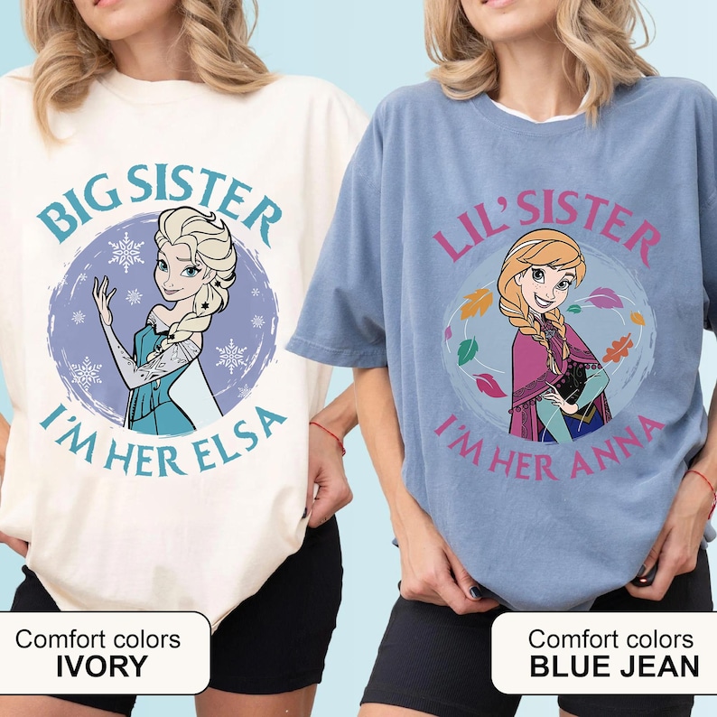 May include: Two t-shirts, one ivory and one blue jean, featuring cartoon images of Elsa and Anna from Frozen. The ivory shirt says "Big Sister, I'm Her Elsa." The blue jean shirt says "Lil' Sister, I'm Her Anna."