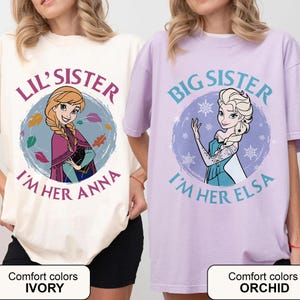 May include: Two t-shirts, one ivory and one orchid, featuring cartoon characters. The ivory shirt says "Lil' Sister, I'm Her Anna" with a picture of Anna. The orchid shirt says "Big Sister, I'm Her Elsa" with a picture of Elsa.