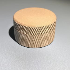 May include: A cylindrical container with a flat top and a textured, diamond-patterned knurled grip around the side. The object is a solid, light beige colour with a matte finish, featuring a clear seam where the lid meets the base.