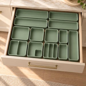 Peut inclure: Un ensemble d'organisateurs de tiroir vert sauge disposés à l'intérieur d'un tiroir de couleur claire. La collection comprend divers plateaux rectangulaires et carrés, certains avec des séparateurs internes, pour trier les objets.