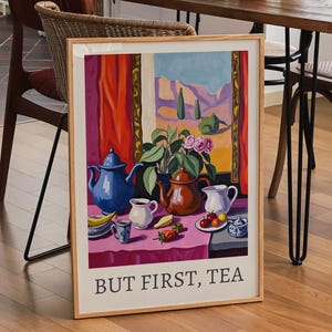 May include: A framed art print with a still life of teapots, cups, and fruit, with the text "BUT FIRST, TEA". The artwork is in a room with a wooden table, chairs, and a bottle of wine. The room has a plant and a window.