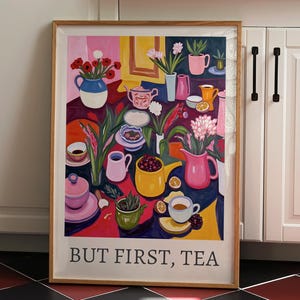 May include: A framed art print with a colorful illustration of a tea party scene. The artwork features various teacups, teapots, and vases filled with flowers, with the text "BUT FIRST, TEA" at the bottom. The color palette includes pinks, blues, yellows, and reds.