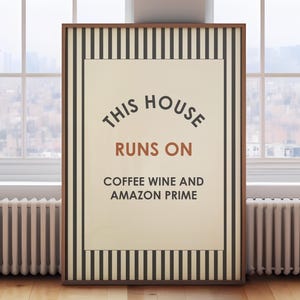 May include: Framed wall art with a cream background and the text "THIS HOUSE RUNS ON COFFEE WINE AND AMAZON PRIME." The frame has a brown border with black and white vertical stripes. The artwork is displayed on a wooden floor.