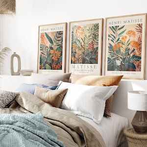 May include: Three framed Henri Matisse art prints with botanical illustrations in orange, green, and brown tones. The prints are displayed above a bed with neutral-colored bedding and pillows. A lamp with a textured base sits on a woven side table.
