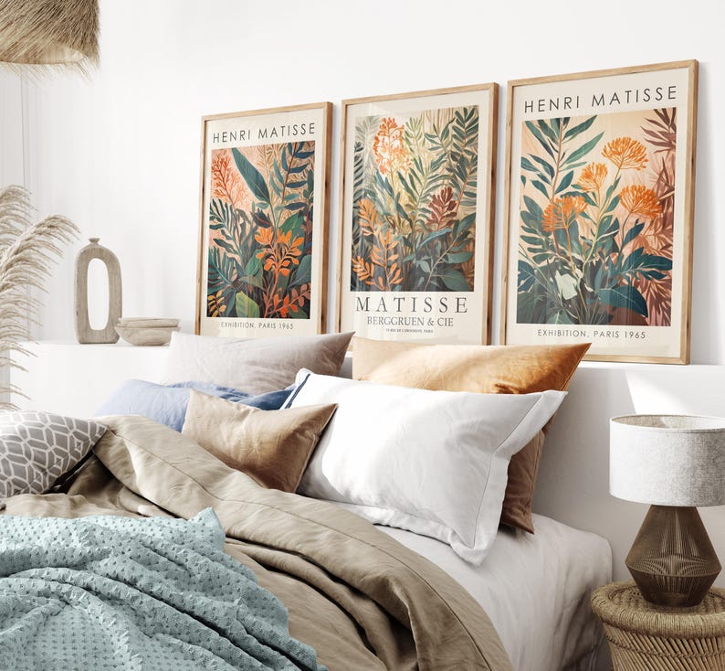 May include: Three framed Henri Matisse art prints with botanical illustrations in orange, green, and brown tones. The prints are displayed above a bed with neutral-colored bedding and pillows. A lamp with a textured base sits on a woven side table.
