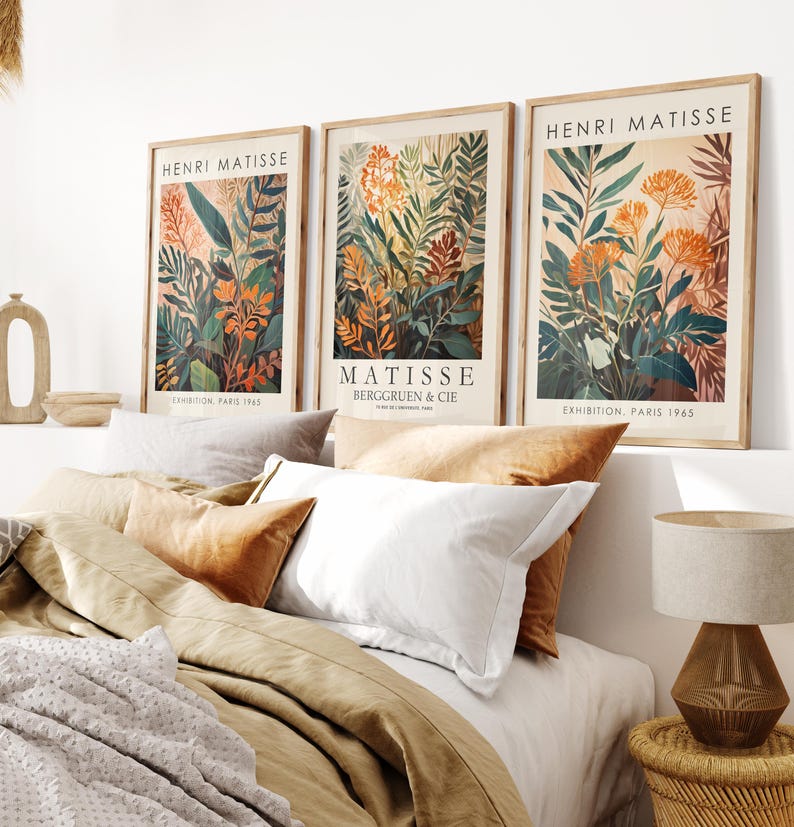 May include: Three framed art prints featuring botanical illustrations in shades of orange, green, and brown. The prints are labeled "HENRI MATISSE" and "MATISSE BERGGRUEN & CIE" with "EXHIBITION, PARIS 1965". The prints are displayed above a bed with pillows and a lamp.