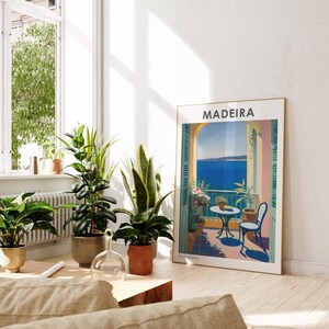 May include: A vintage travel poster with the word "MADEIRA" in bold white letters on a blue background. The poster depicts a balcony overlooking a blue ocean with a table and chairs on the balcony. The balcony is surrounded by lush green plants.