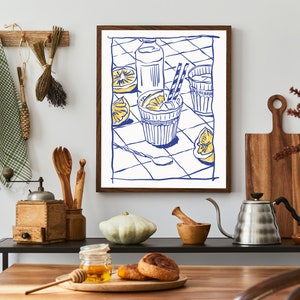 Pu&ograve; includere: Una stampa artistica incorniciata raffigura un'illustrazione blu e bianca di una bevanda rinfrescante con limoni. L'opera d'arte &egrave; esposta in un ambiente cucina, insieme a utensili di legno, un macinacaff&egrave; e un tagliere, creando un'atmosfera accogliente.