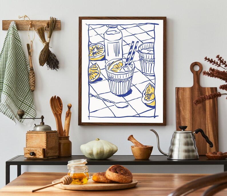 Pu&ograve; includere: Una stampa artistica incorniciata raffigura un'illustrazione blu e bianca di una bevanda rinfrescante con limoni. L'opera d'arte &egrave; esposta in un ambiente cucina, insieme a utensili di legno, un macinacaff&egrave; e un tagliere, creando un'atmosfera accogliente.