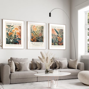 May include: Three framed Henri Matisse art prints featuring botanical illustrations in orange, green, and beige tones. The prints are displayed above a gray sofa with decorative pillows. The room has a modern, minimalist design.