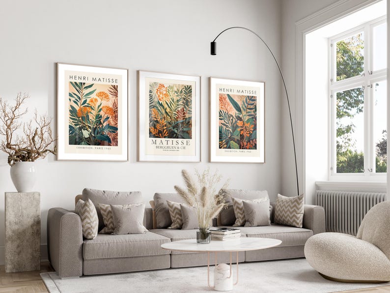 May include: Three framed Henri Matisse art prints featuring botanical illustrations in orange, green, and beige tones. The prints are displayed above a gray sofa with decorative pillows. The room has a modern, minimalist design.