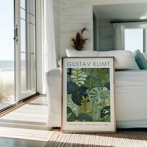May include: A framed art print featuring a botanical illustration in shades of green and blue. The print is labeled "GUSTAV KLIMT" and "EXHIBITION, AMSTERDAM 1988". The artwork is displayed on a white couch in a bright room with a view of the ocean.