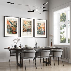 May include: Three framed Henri Matisse botanical art prints with black frames hang above a dining table. The prints feature colorful floral and plant illustrations. A modern dining table with gray chairs and a black light fixture complete the scene.