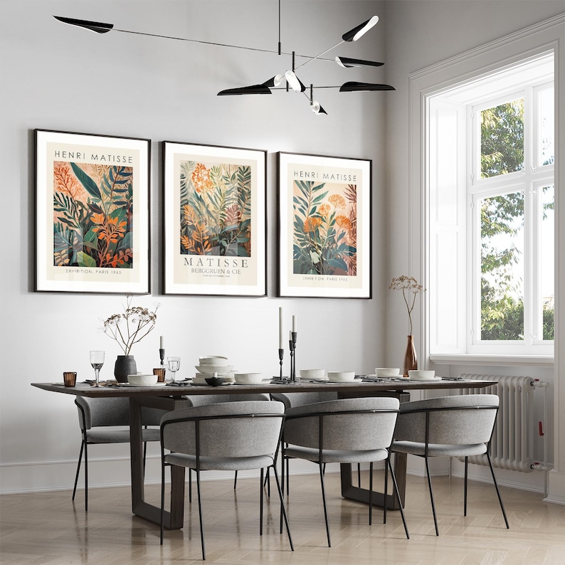 May include: Three framed Henri Matisse botanical art prints with black frames hang above a dining table. The prints feature colorful floral and plant illustrations. A modern dining table with gray chairs and a black light fixture complete the scene.