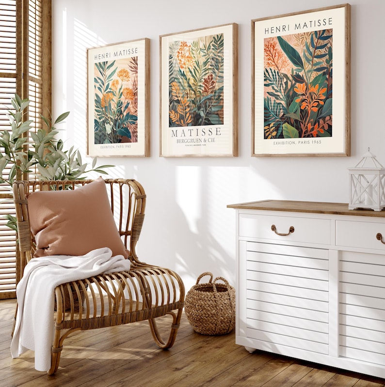 May include: Three framed Henri Matisse art prints featuring botanical illustrations in shades of green, orange, and beige. The prints are displayed above a white cabinet and a wicker chair with a pillow and blanket. A woven basket sits on the floor.