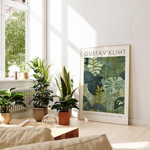 May include: A framed art print featuring a Gustav Klimt botanical illustration in shades of green and blue. The print is displayed in a bright room with several potted plants. Text on the print reads "GUSTAV KLIMT" and "EXHIBITION, AMSTERDAM 1988."