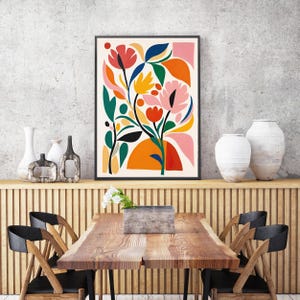 May include: A framed floral art print with a black border. The artwork features a colorful abstract design of flowers and leaves in shades of red, pink, orange, yellow, green, and blue against a cream background. The print is displayed above a wooden table.
