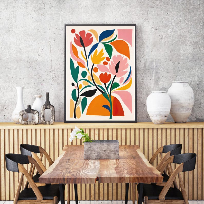 May include: A framed floral art print with a black border. The artwork features a colorful abstract design of flowers and leaves in shades of red, pink, orange, yellow, green, and blue against a cream background. The print is displayed above a wooden table.