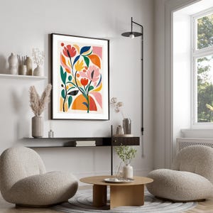 May include: A framed floral art print with a black border. The artwork features abstract flowers in shades of pink, orange, yellow, and blue against a cream background. The print is displayed in a modern living room setting.