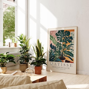Puede incluir: Un cuadro enmarcado con una ilustraci&oacute;n bot&aacute;nica de varias plantas y hojas verdes en macetas. El cuadro est&aacute; apoyado contra una pared blanca, junto a varias plantas en macetas. El texto "MATISSE BERGGRUEN & CIE" es visible.