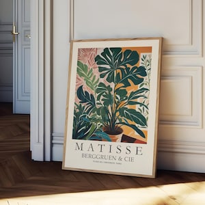 Puede incluir: Un grabado art&iacute;stico enmarcado con una ilustraci&oacute;n bot&aacute;nica de varias plantas verdes en una maceta de terracota. La obra de arte tiene un fondo beige con el texto "MATISSE BERGGRUEN & CIE" y la direcci&oacute;n "70 RUE DE L'UNIVERSITE, PARIS". El marco es de madera marr&oacute;n claro.