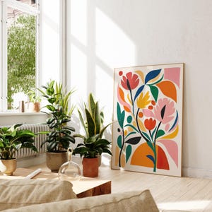 May include: A framed floral art print with a colorful abstract design. The artwork features pink, red, yellow, and blue flowers and leaves against a cream background. The print is displayed in a bright, sunlit room with potted plants.